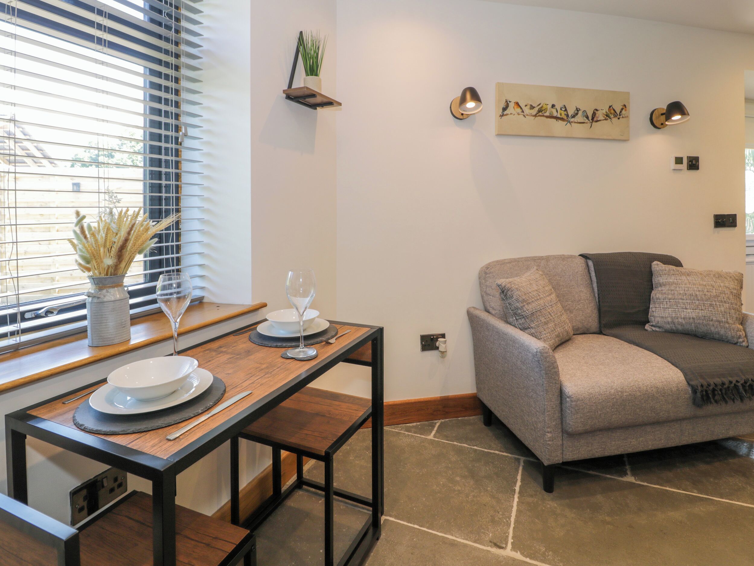 A modern living room with a dining area, featuring a sofa, a table with plates and a wine glass, and wall-mounted lamps.