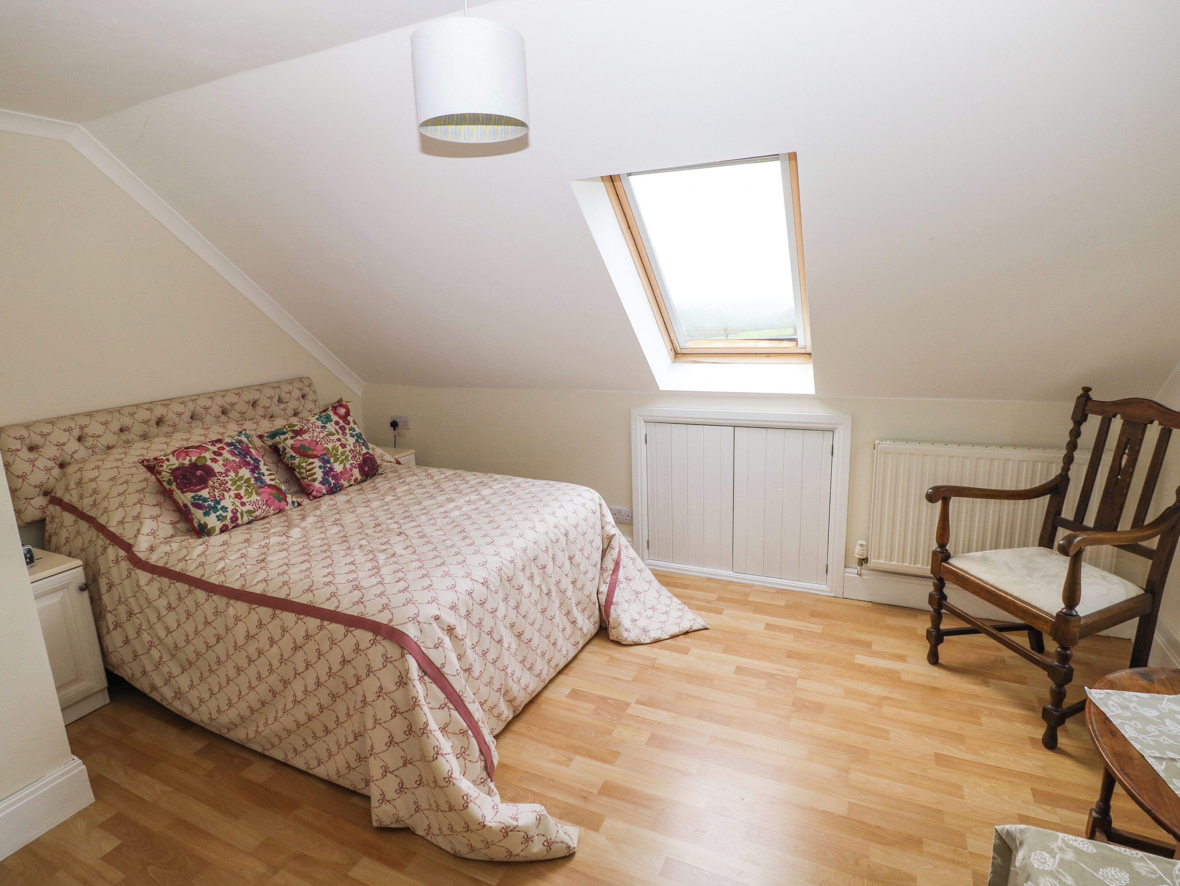 A bedroom with a sloped ceiling, a skylight, a bed with patterned bedding, a wooden chair, and a radiator.