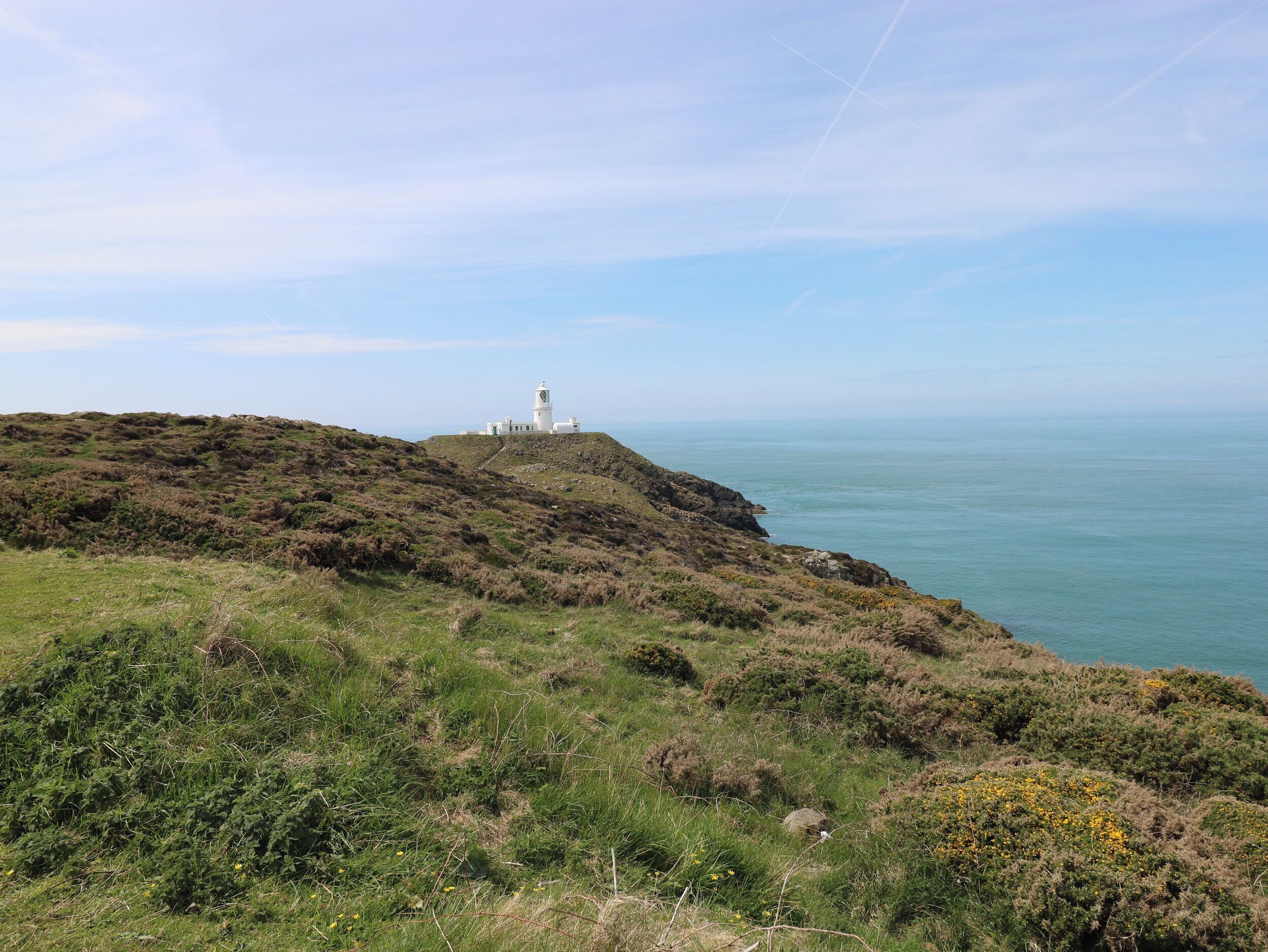 A coastal landscape with a white lighthouse on a grassy hill overlooking the sea.