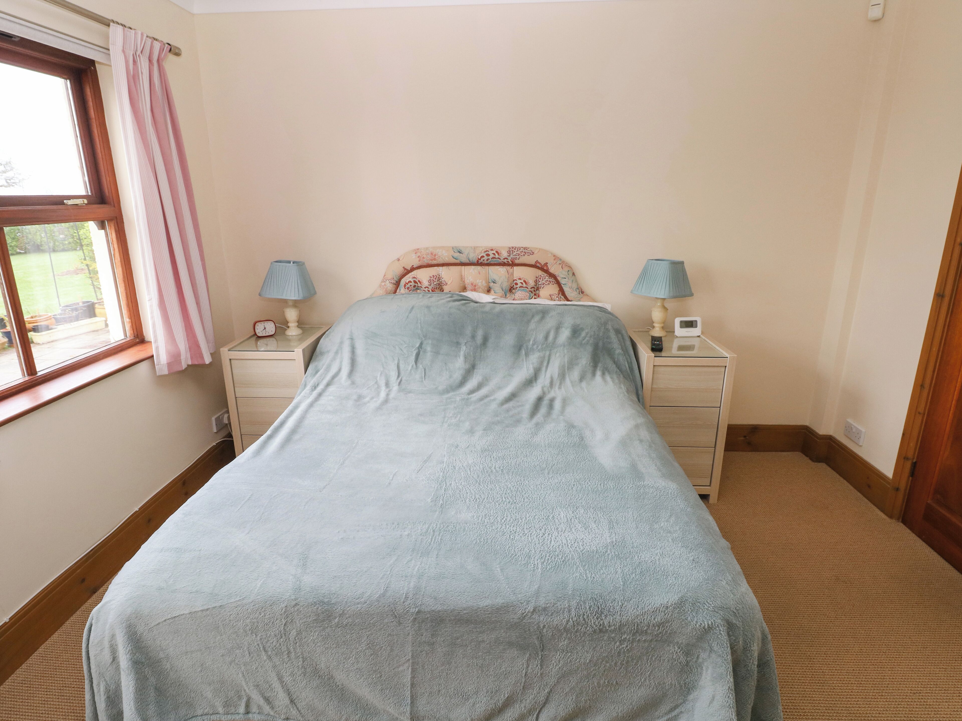 A bedroom with a bed, two bedside tables with lamps, a window with curtains, and a view of the outdoors.