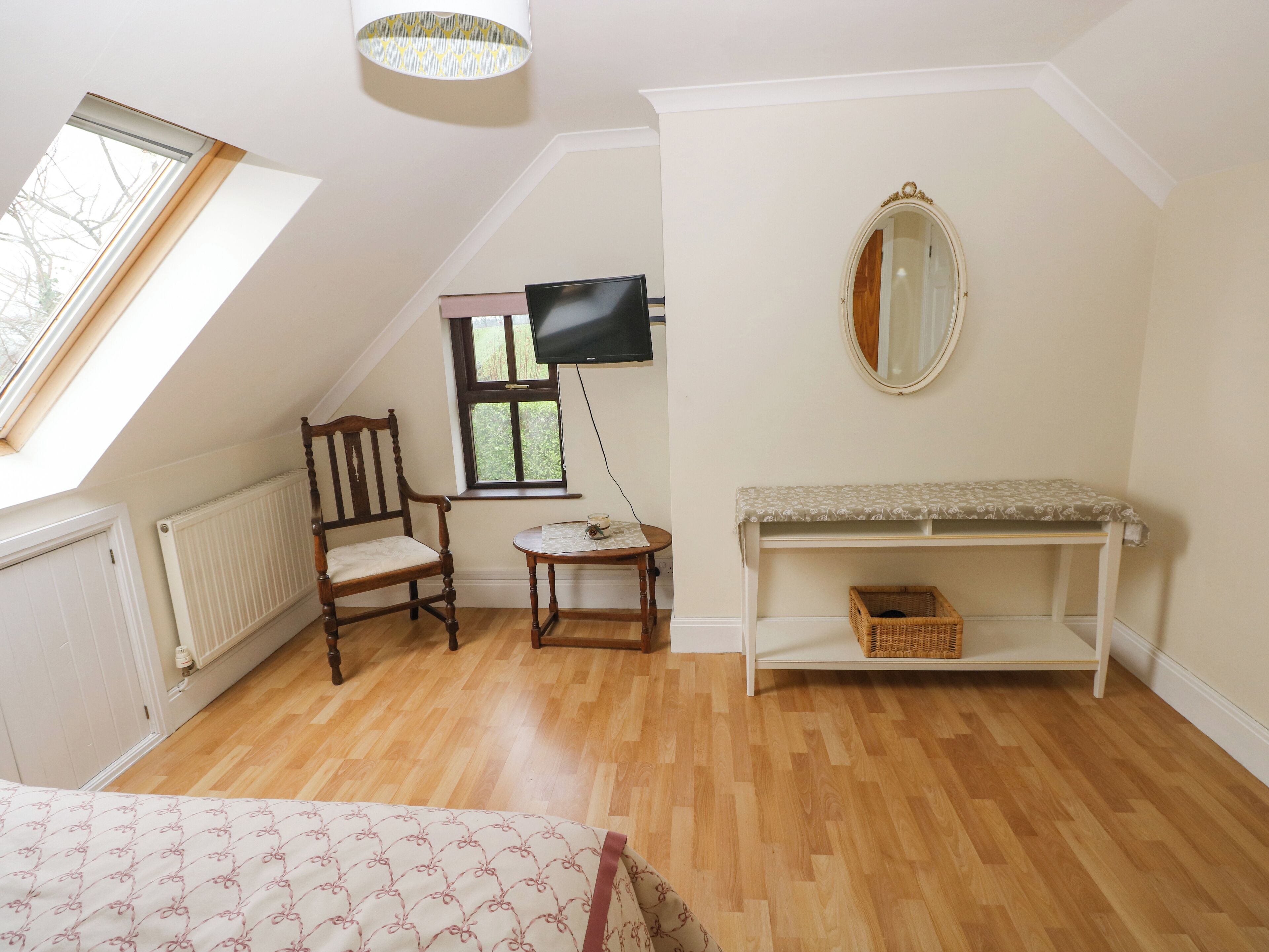 A room with a sloped ceiling, wooden flooring, a bed, a chair, a small table, a TV, a mirror, and a bench with a basket.