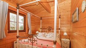 A four-poster bed with a wooden headboard, a bedside table with a lamp, and a window with curtains. - Woodpecker Lodge (Bury St Edmunds)