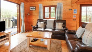 A cozy cabin interior with a wooden ceiling, leather sofas, a wooden coffee table, and a TV stand. - Woodpecker Lodge (Bury St Edmunds)