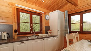 A wooden cabin kitchen with a sink, a kettle, a bread container, and a dining table with a polka dot tablecloth. - Woodpecker Lodge (Bury St Edmunds)