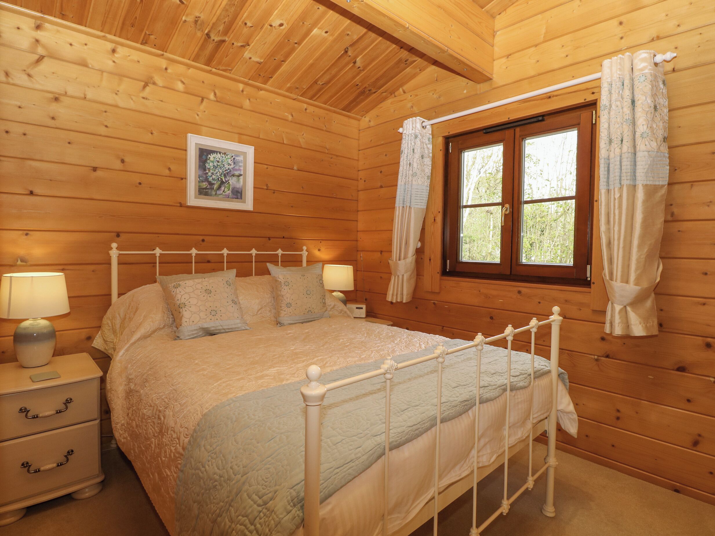 A wooden cabin bedroom with a bed, bedside lamps, a window with curtains, and a framed picture on the wall.