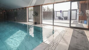 An indoor swimming pool with clear water, a stone wall, and a glass door leading to an outdoor seating area. - Lodge 3 (Saint Asaph)