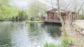 A wooden cabin with a deck overlooking a calm river, surrounded by lush greenery and trees. - Kingfisher Lodge (Bury St Edmunds)