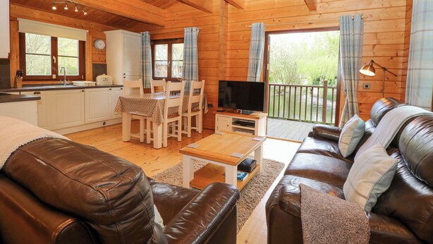 A cozy cabin interior with a dining area, a living room with leather sofas, a television, and a kitchen with a window overlooking a garden. - Kingfisher Lodge (Bury St Edmunds)