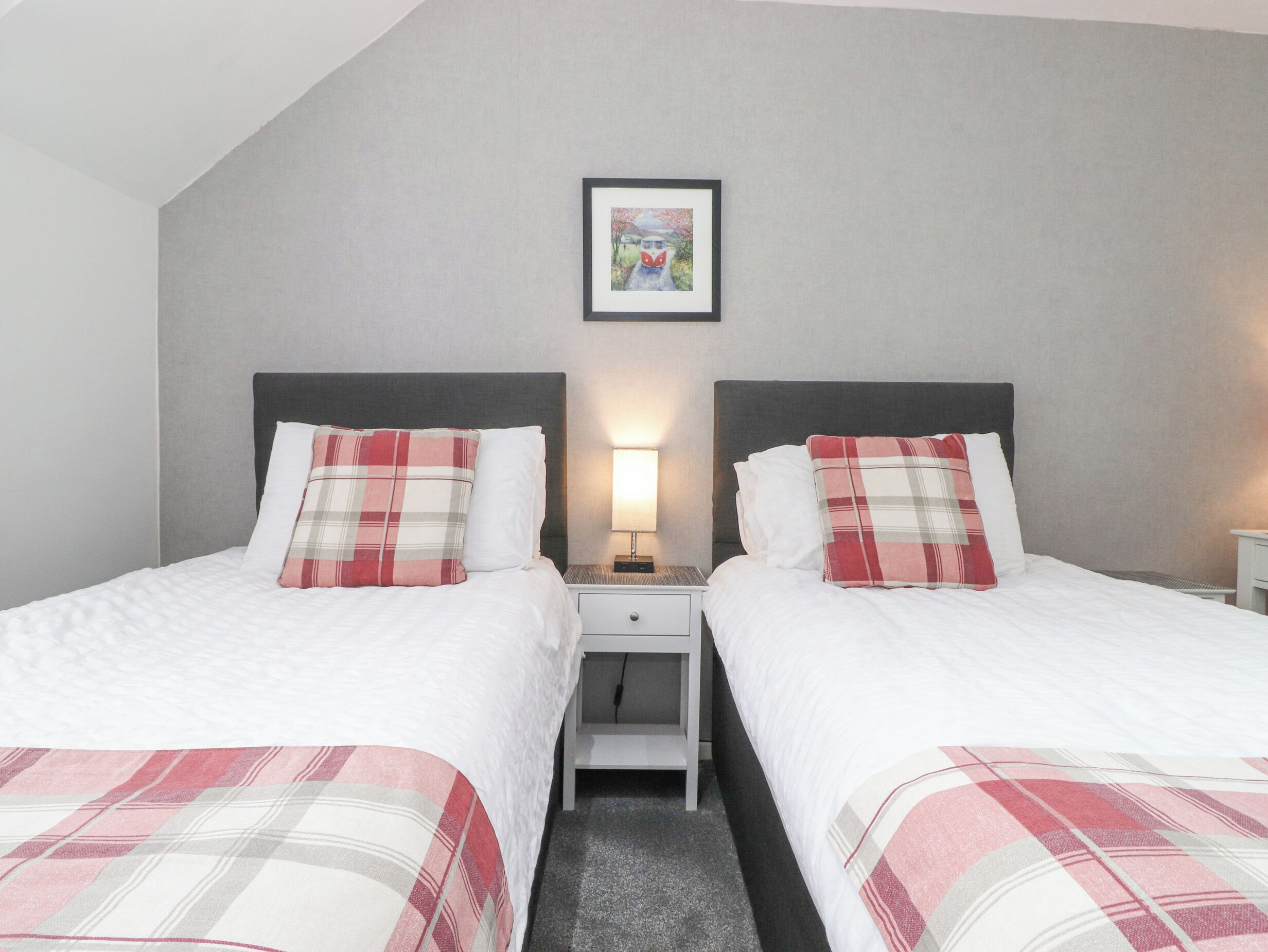 A hotel room with two beds, each with a plaid bedspread and pillows, a nightstand with a lamp, and a framed picture on the wall.