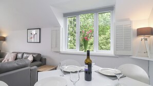 A modern living room with a dining area, a grey sofa, a white table set for four, and a window with a view of greenery. - The Penthouse (Windermere)