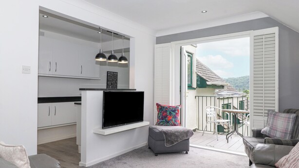 A modern kitchen with white cabinets, a black countertop, and a TV. There is a grey sofa with a patterned throw pillow, a small grey ottoman, and a white chair. A balcony with a view of a house and trees is visible. - The Penthouse (Windermere)