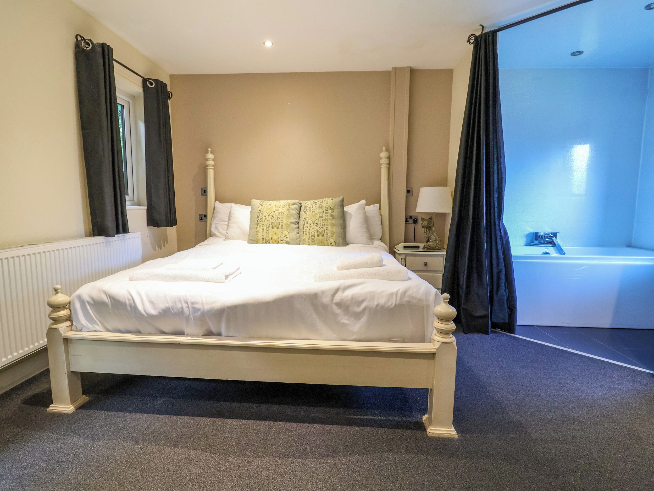 Un lit bien fait, avec du linge de lit blanc, une table de chevet avec une lampe, et une vue sur une salle de bain avec une baignoire.