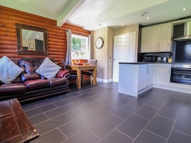 A log cabin interior with a leather sofa, wooden dining table, and a kitchen area with white cabinets and a built-in oven.