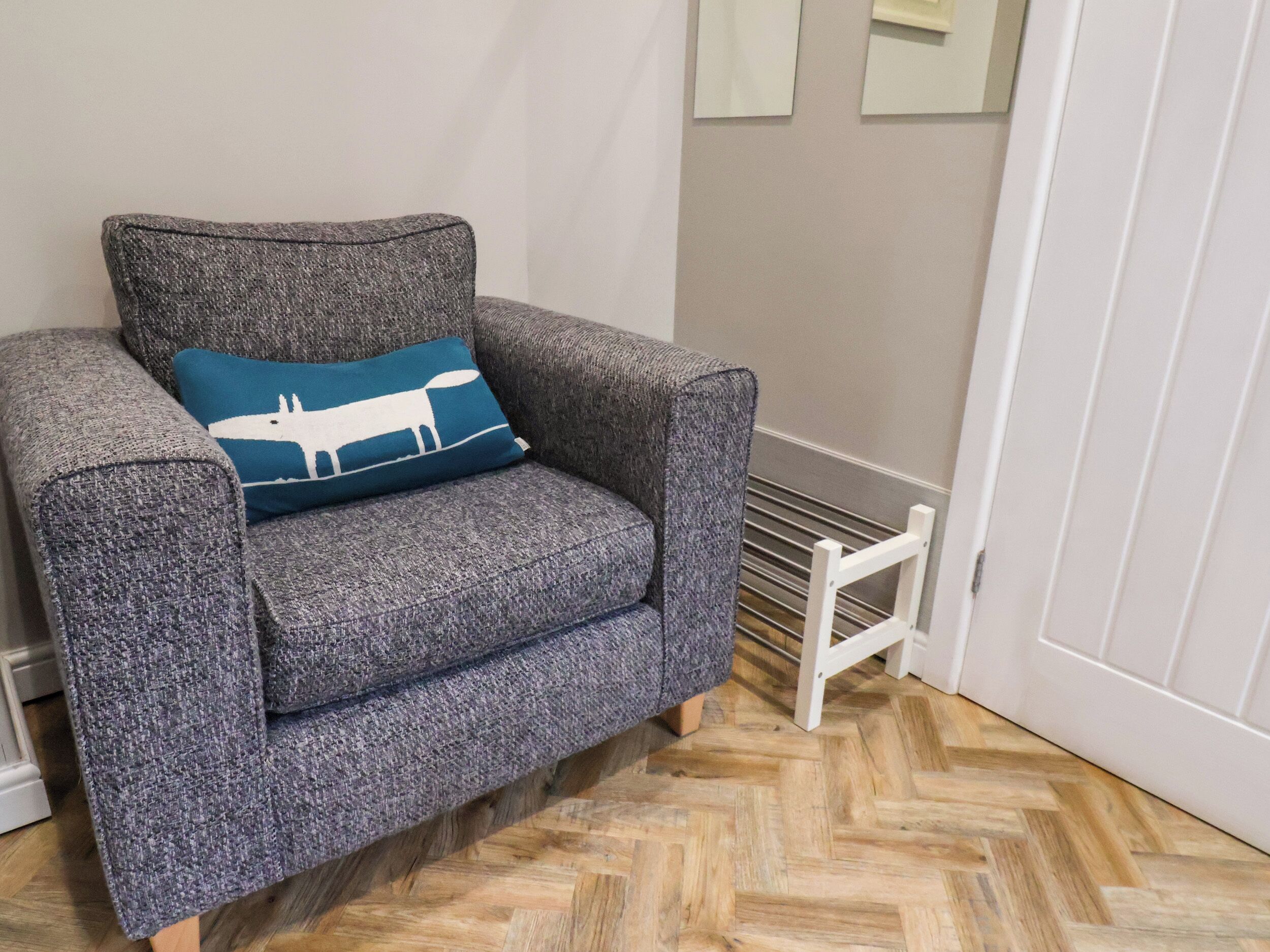 Un fauteuil gris avec un coussin bleu orné de la silhouette blanche d’un animal, placé près d’un radiateur blanc et d’un parquet en bois à motifs chevrons.