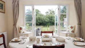 A dining room with a table set for four, two chairs, a window with curtains, and a view of a garden. - Culver Court Cottage (Crediton)