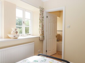 A room with a window, a radiator, a door leading to a bathroom, and a bed with a floral bedspread. - Culver Court Cottage (Crediton)