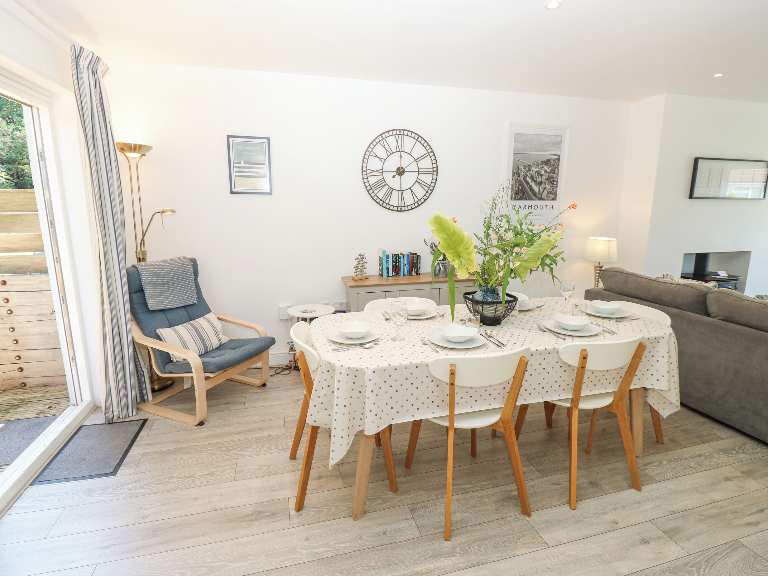 Uma sala de jantar com uma mesa redonda posta para seis pessoas, rodeada por cadeiras. Há um relógio na parede, uma moldura com uma fotografia e um vaso com flores sobre a mesa.