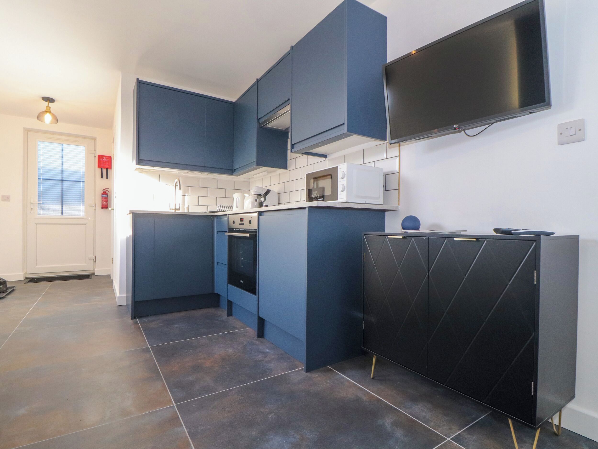 Een moderne keuken met blauwe kastjes, een zwarte koelkast, een magnetron en een flatscreen televisie aan de muur.