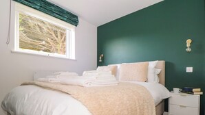 A neatly made bed with folded towels, a bedside table with a cup and books, and a window with a view of trees. - Fifty Degrees North (Penzance)