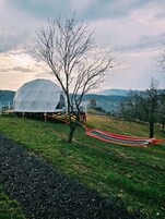 Estructura en forma de cúpula geométrica con una terraza de madera y una hamaca en un campo cubierto de hierba, con árboles y colinas al fondo.