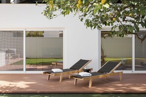 A modern outdoor lounge area with two sun loungers, a wooden deck, and a glass wall offering a view of a garden and a pool. - Athena's Lemon Grove Glyfada (Voula)
