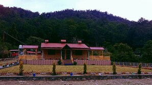 A lodge with a central wooden structure, a red roof, and a stone foundation. There are multiple rooms with balconies, and the property is surrounded by a well-maintained garden with a variety of plants and a stone pathway leading to the entrance. - River View Bamboo Resort (Lansdowne)