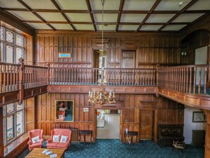 A spacious room with wooden paneling, a chandelier, a piano, and a balcony with a railing. - Alston Hall (Plymouth)