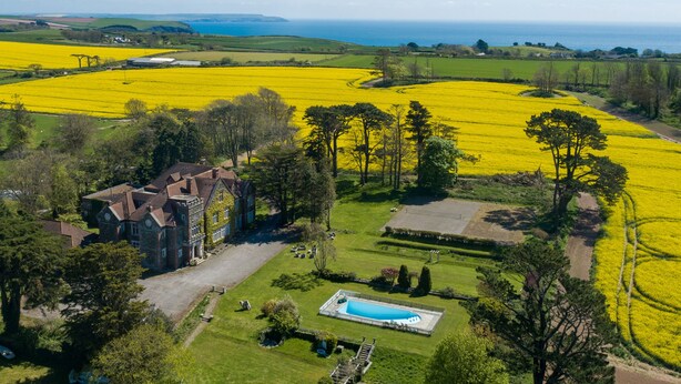 Vista aérea de una extensa propiedad con piscina, rodeada de amplios campos de flores amarillas y una casa grande de estilo arquitectónico tradicional.