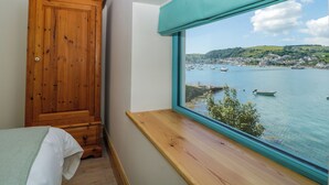 A bedroom with a large window overlooking a harbor, a wooden wardrobe, and a bed with a white cover. - Britannia Crossings Cottage (Dartmouth)
