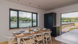 A dining area with a wooden table set for six, white chairs, and a view of a lake with boats. - Britannia Crossings Cottage (Dartmouth)