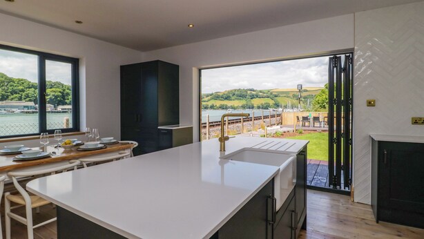 A modern kitchen with a large island, a view of the outdoors, and a dining area with chairs and a table. - Britannia Crossings Cottage (Dartmouth)