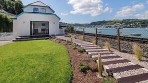 A modern house with a white exterior and blue trim, a small front yard with a lawn and stepping stones, and a view of a harbor with boats and a town in the background. - Britannia Crossings Cottage (Dartmouth)
