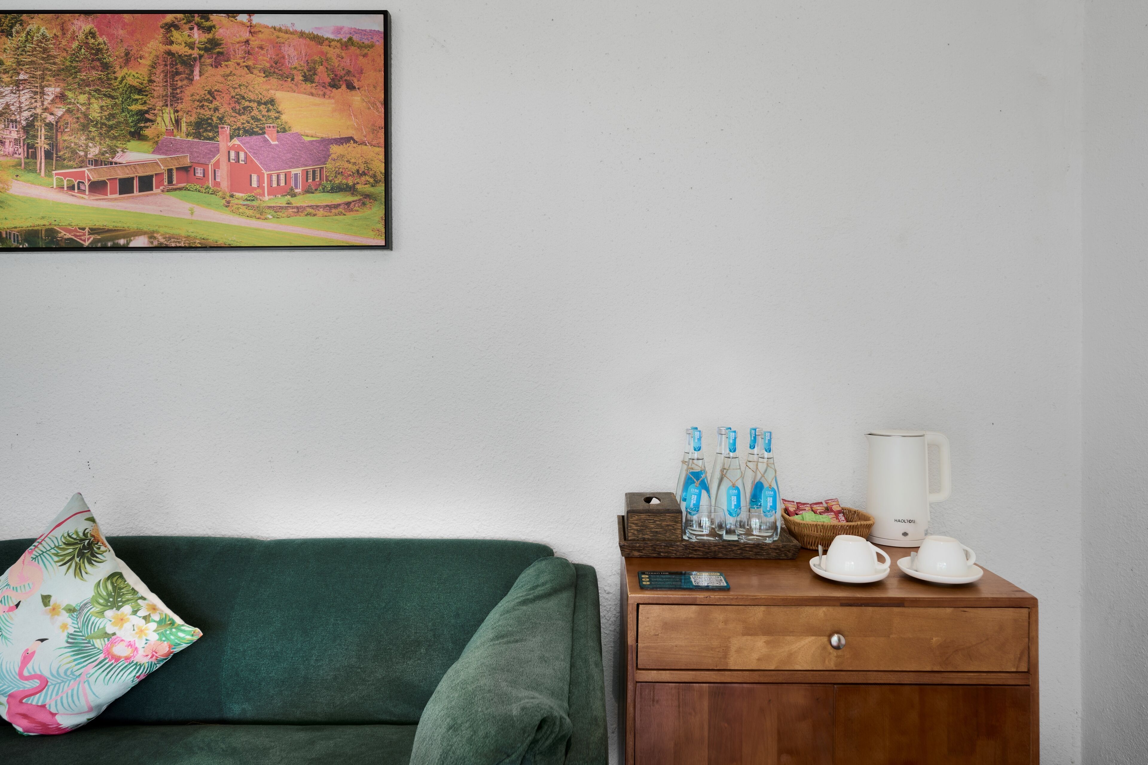 A room with a green sofa, a wooden nightstand with a kettle, and a painting of a house on the wall.