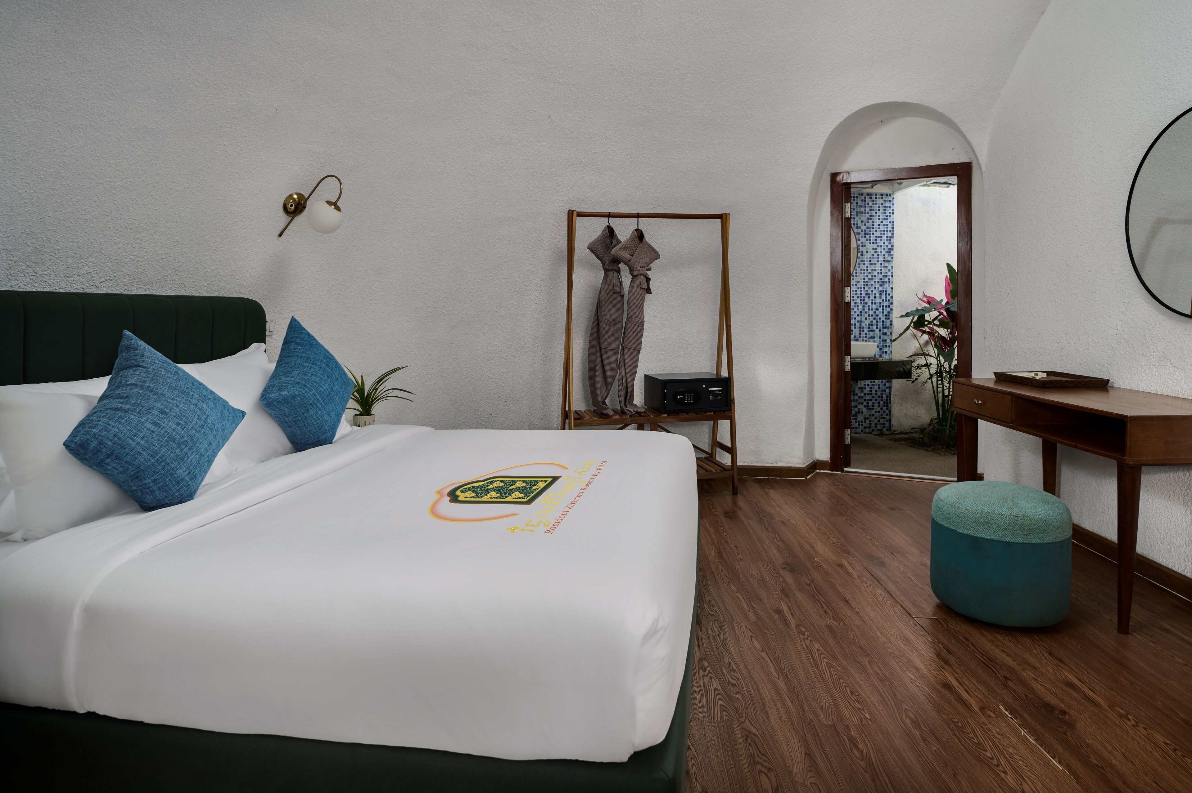 A hotel room with a neatly made bed, a small desk, a mirror, and a wooden floor.