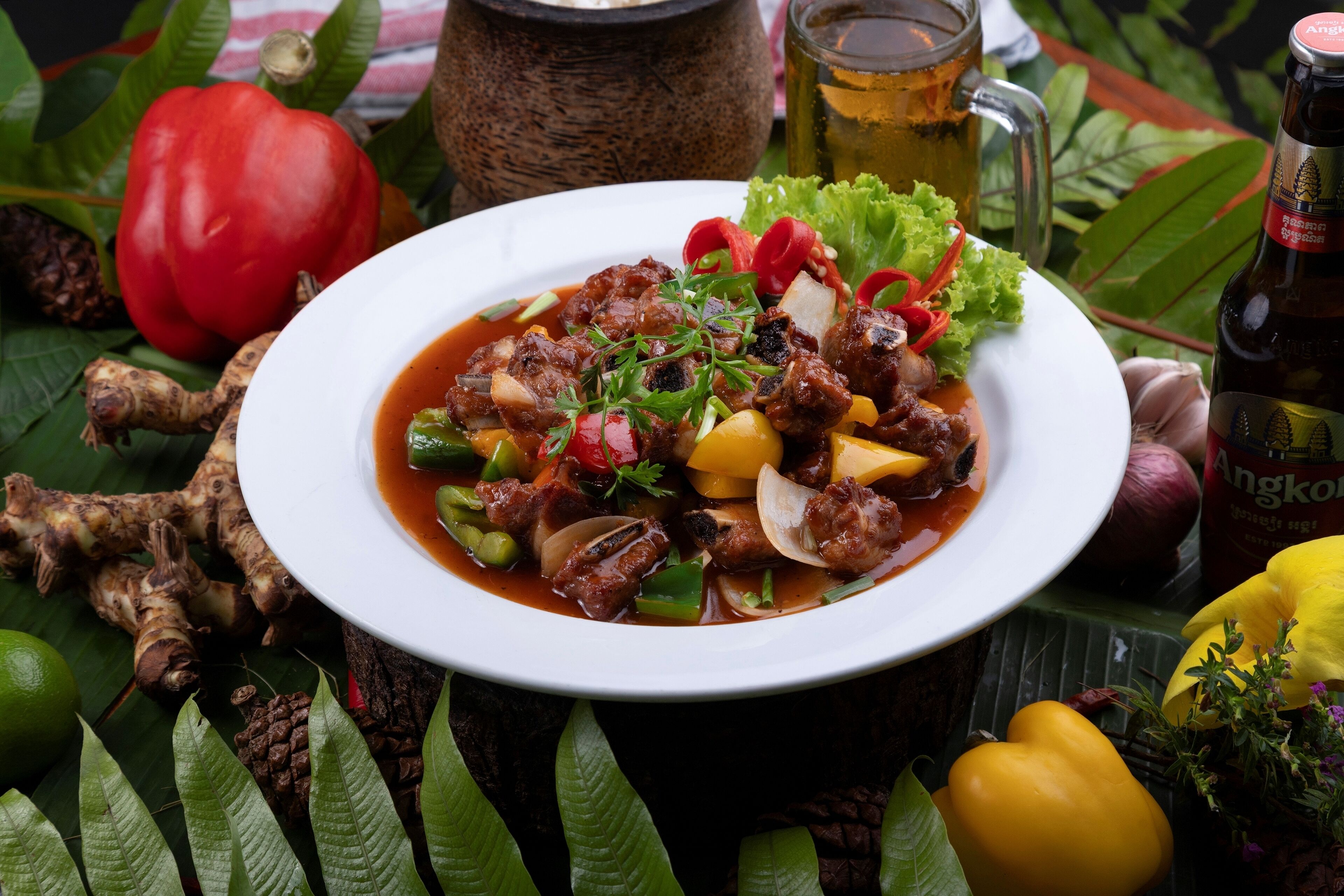Un tazón de guiso con verduras y carne, rodeado de productos frescos y una botella de cerveza.