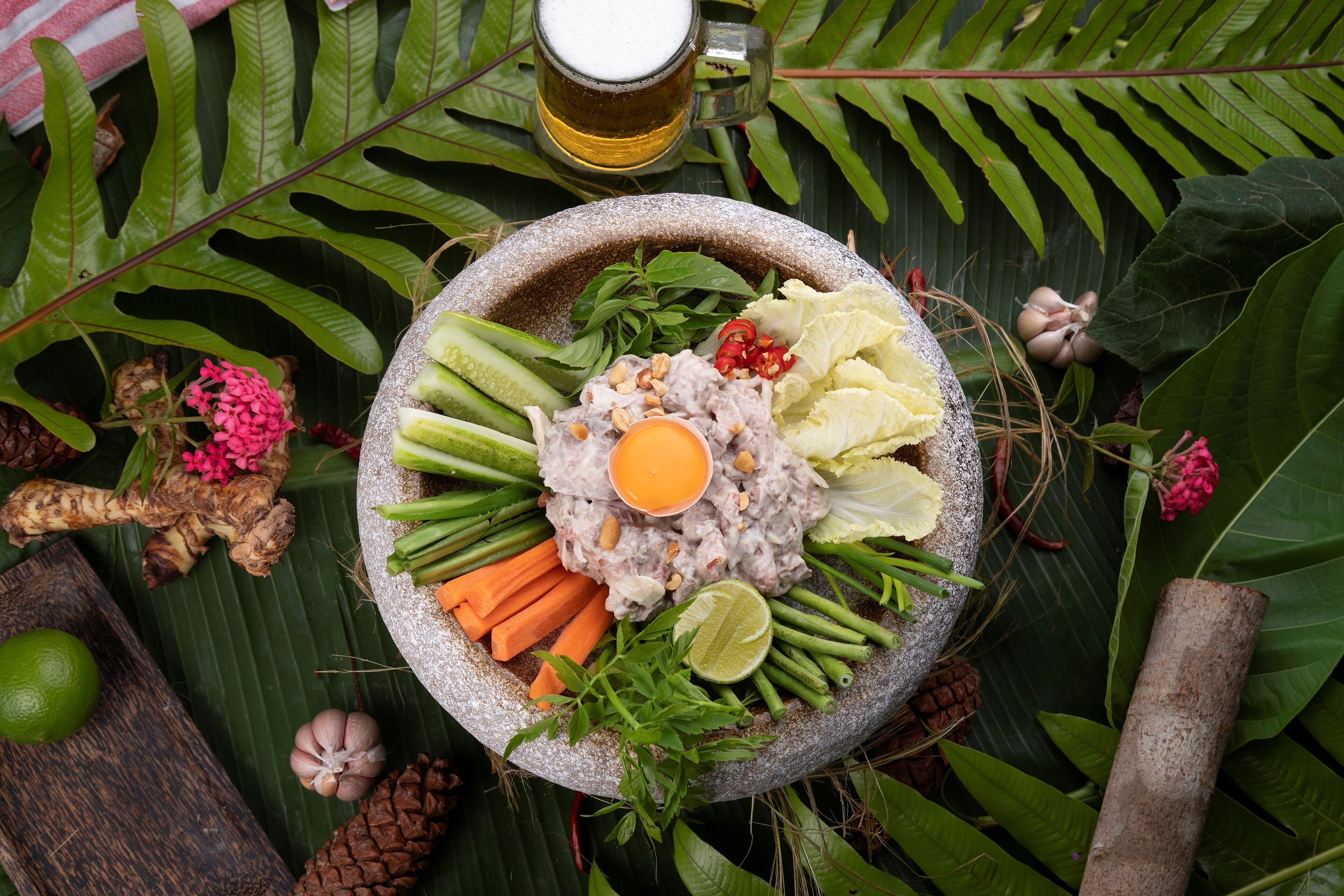 A stone bowl filled with various fresh ingredients like vegetables, herbs, and a lime wedge, surrounded by tropical foliage and a glass of beer.