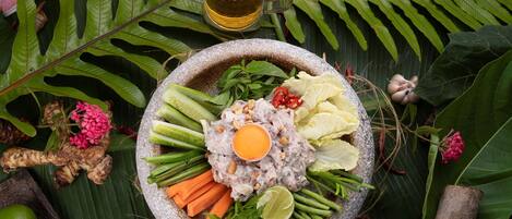 A stone bowl filled with various fresh ingredients like vegetables, herbs, and a lime wedge, surrounded by tropical foliage and a glass of beer.