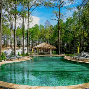 Una piscina rodeada de árboles, con sillas de descanso y una estructura de techo de paja.