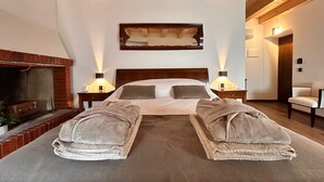 Un dormitorio con techo de madera, chimenea, dos mesitas de noche con lámparas, cama con almohadas y dos toallas sobre la cama.