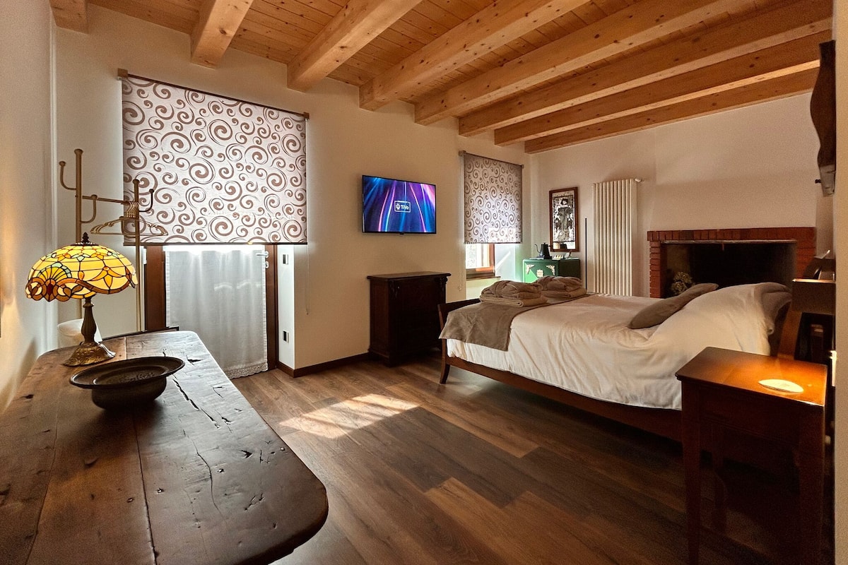 A bedroom with a wooden ceiling, a bed, a TV, a window with curtains, and a bedside table with a lamp.