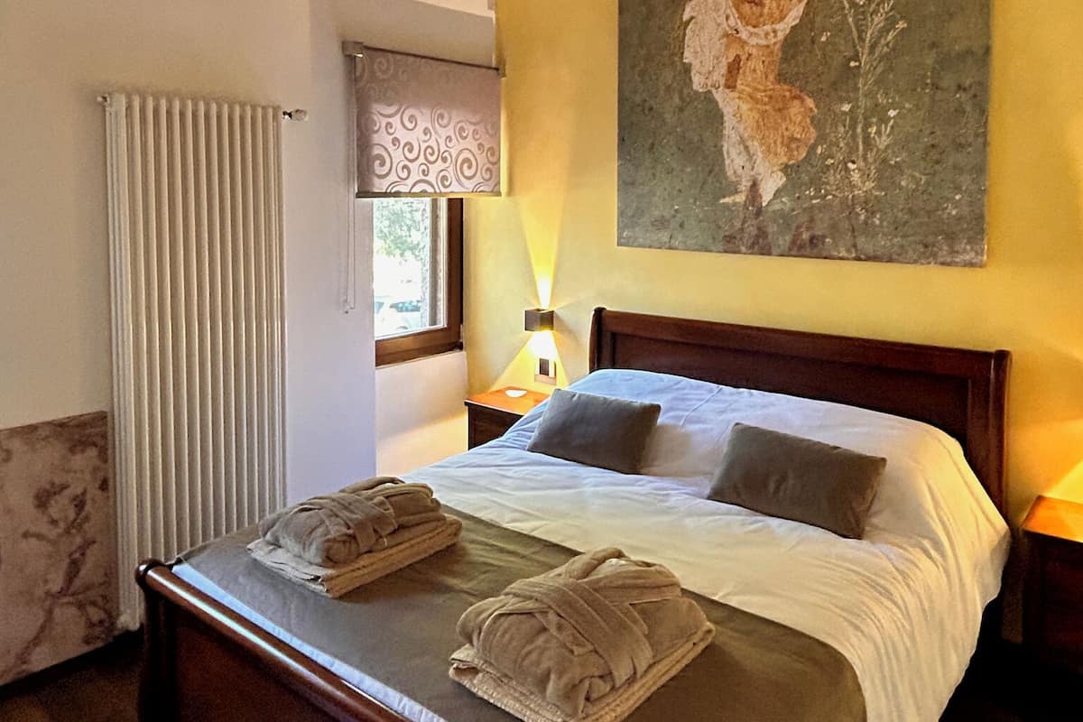 A bedroom with a wooden bed, a radiator, a window with curtains, a wall-mounted painting, and a bedside lamp.