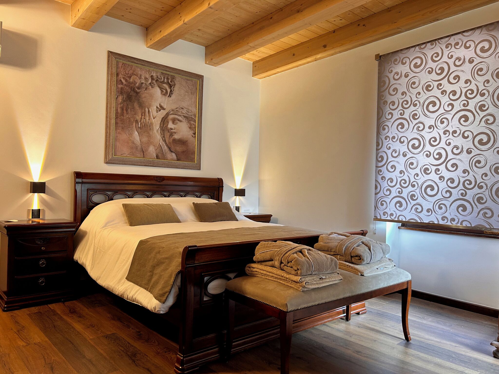 A bedroom with a wooden bed, nightstands, a bench with towels, a large painting, and a patterned curtain.