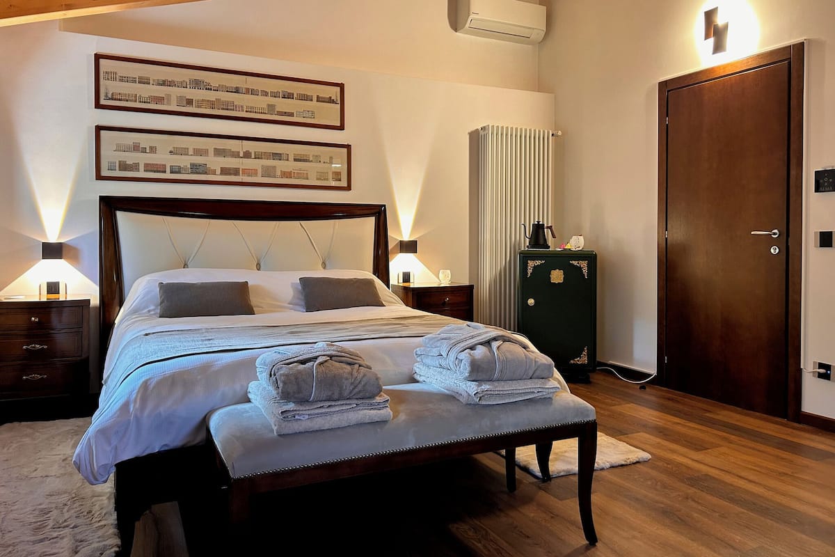 A hotel room with a bed, bedside lamps, a radiator, a wardrobe, and a door with a light switch.