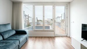A modern living room with a grey sofa, a flat-screen TV, and large sliding glass doors leading to a balcony with a view of the city.