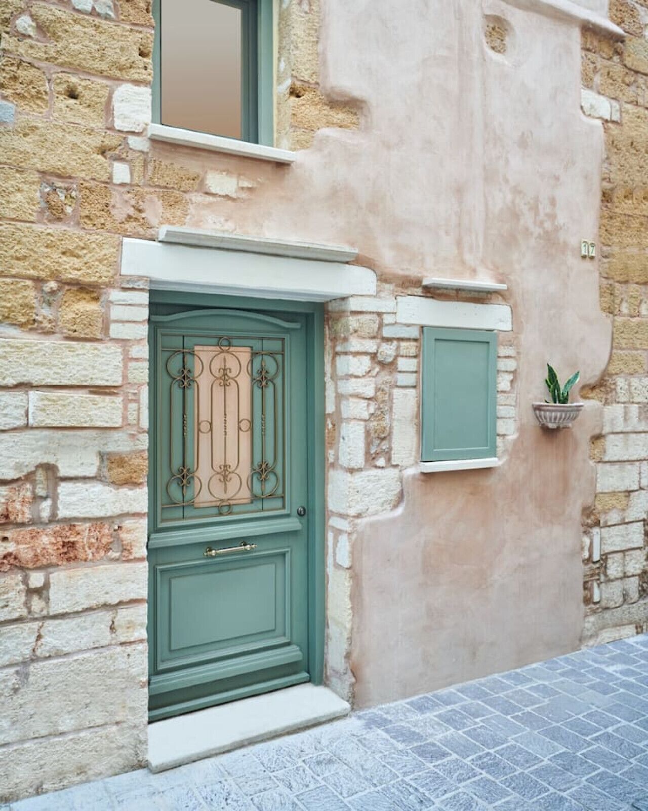 Une façade d’immeuble avec une porte verte, une petite fenêtre au-dessus et un pot de fleurs sur le côté droit.
