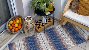 A corner of a room with a white wall, a macrame mirror, a small table with a plant and a pitcher, a basket of oranges, a checkered chessboard, and a sofa with a cushion. - Haplotēs Rhodes Apartment (Koskinou)