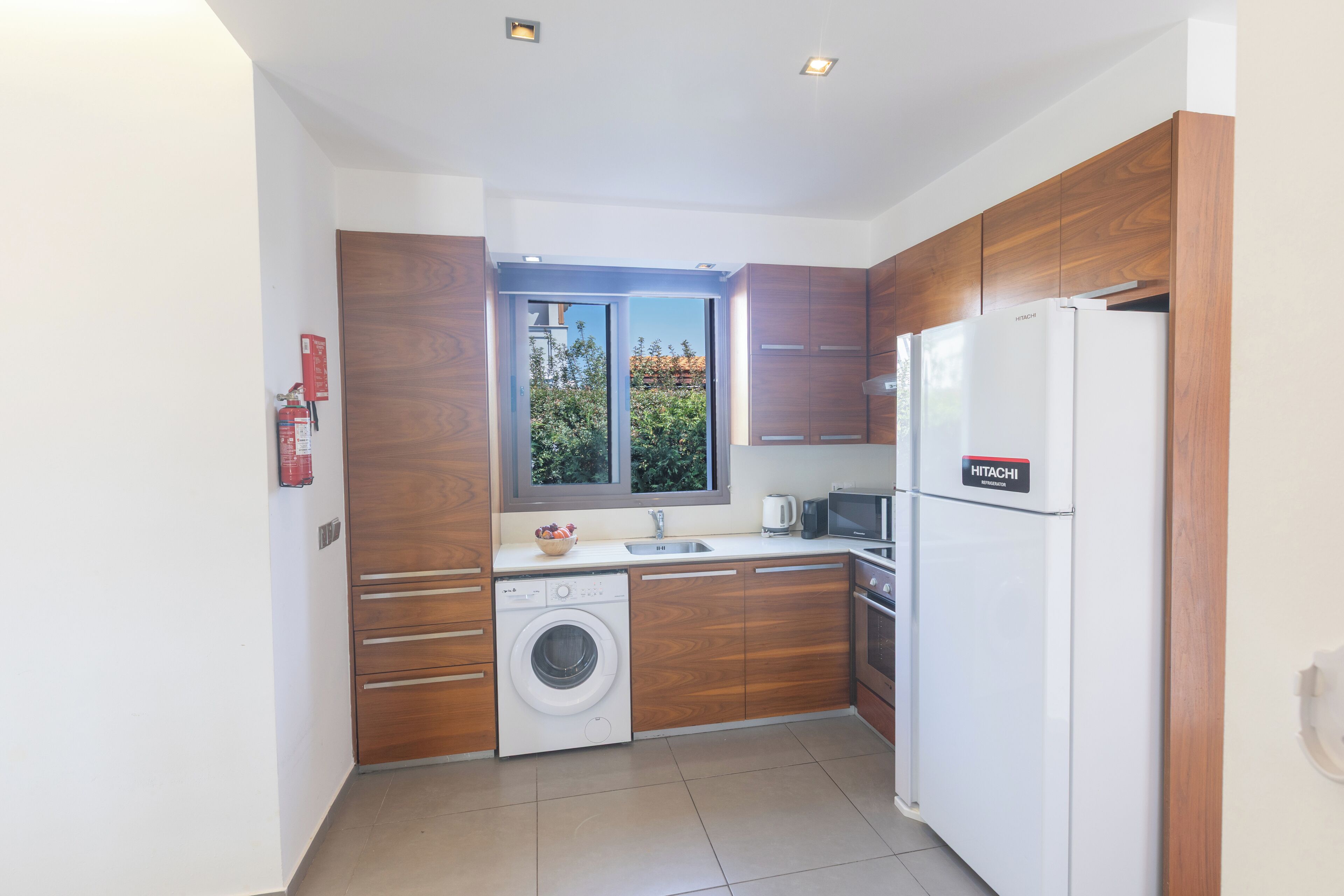 Une cuisine moderne avec des meubles en bois, un lave-linge, un réfrigérateur et une fenêtre donnant sur un paysage verdoyant.