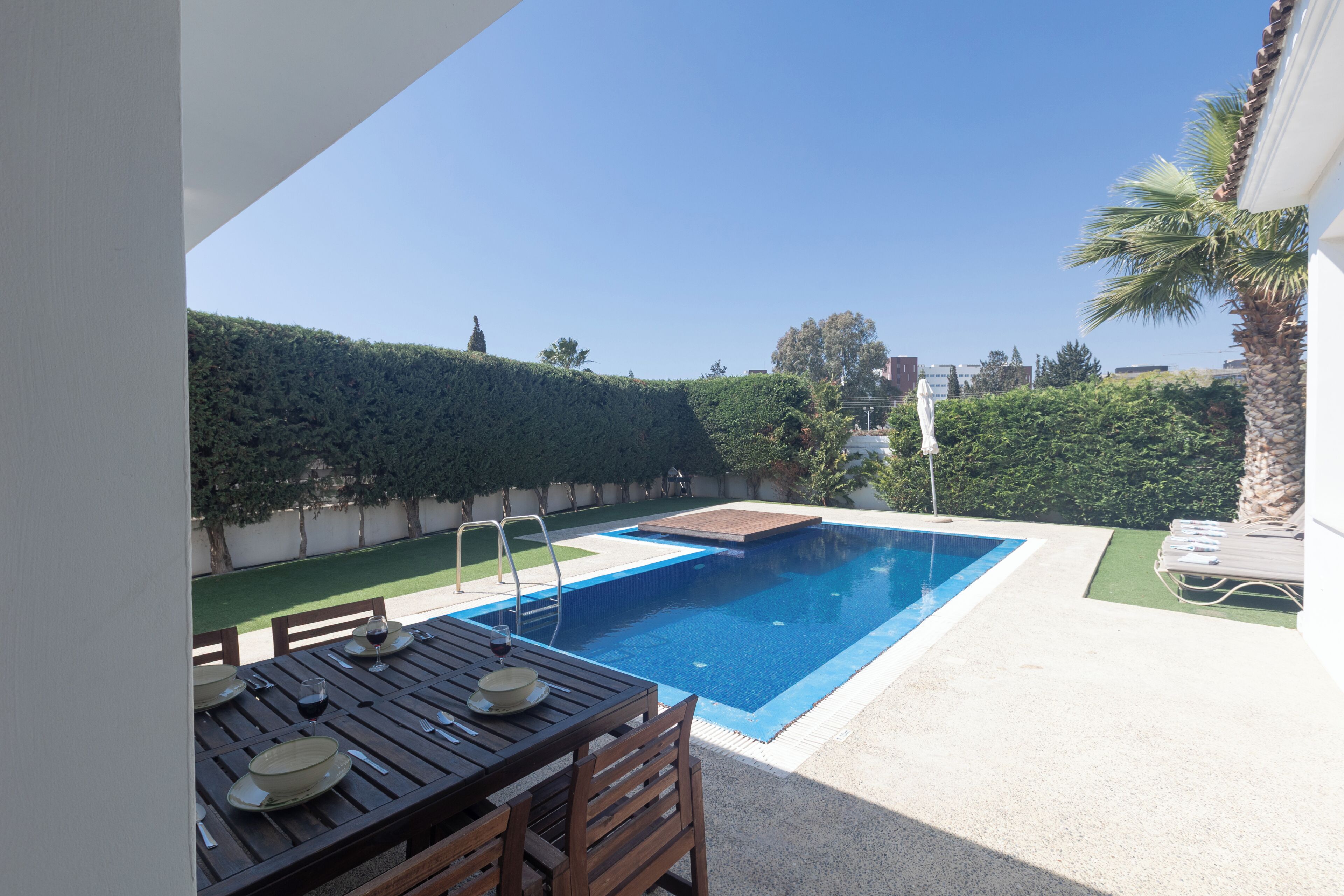 Uma piscina retangular com plataforma de mergulho, mesa de madeira com cadeiras e uma palmeira.