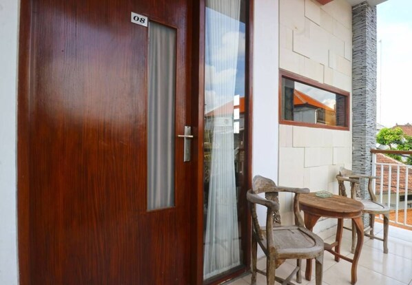 Une porte en bois avec une plaque portant un numéro, une porte-fenêtre coulissante et un petit espace extérieur aménagé avec deux chaises et une table en bois.