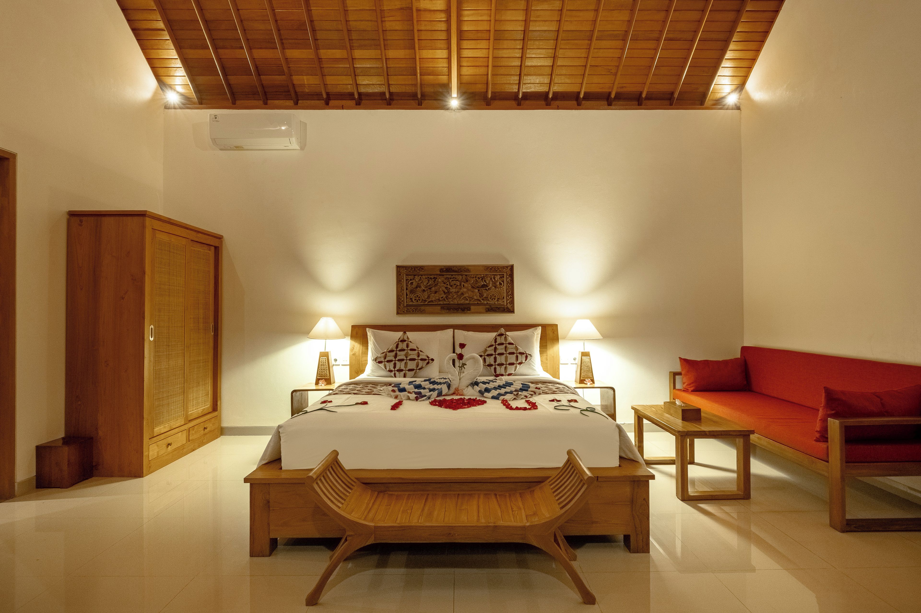 Un dormitorio con una cama, lámparas de noche, un banco de madera, un sofá rojo y un armario de madera.